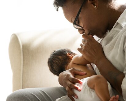 Frau sitzt auf einer Couch, hält ein neugeborenes Baby und küsst die Hand des Babys.