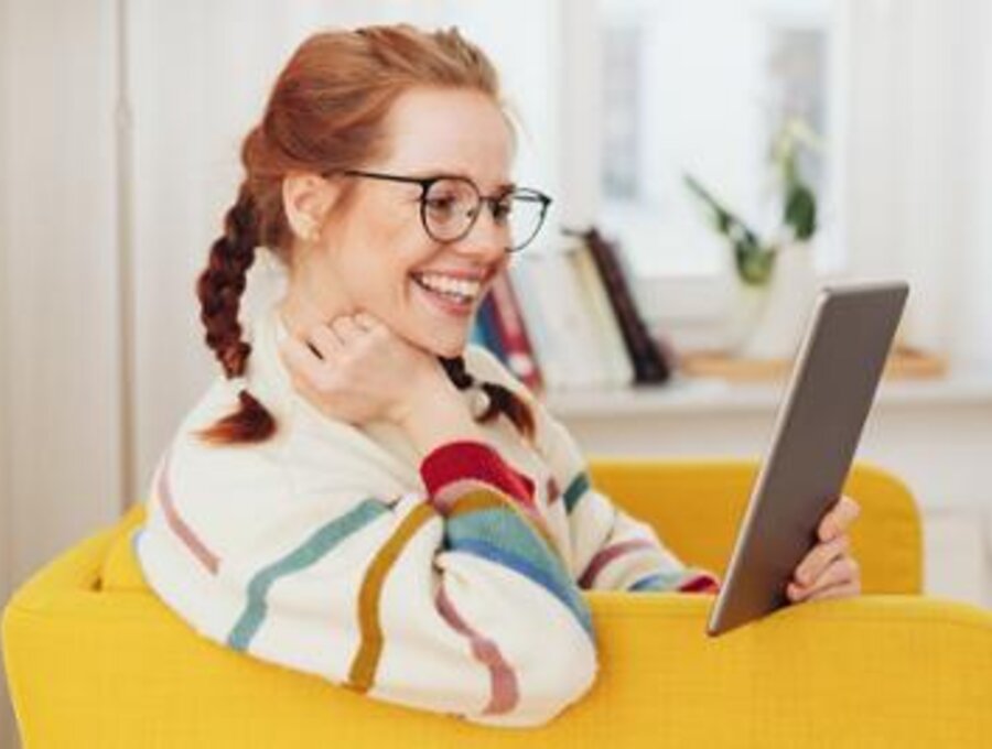 Een vrouw zit thuis op een stoel te lachen en naar haar tablet te kijken.
