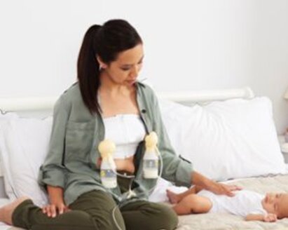 Vrouw zit op bed met een baby naast zich.  Ze kolft dubbel met klassieke borstschilden met een Swing Maxi terwijl ze één hand op de baby legt.