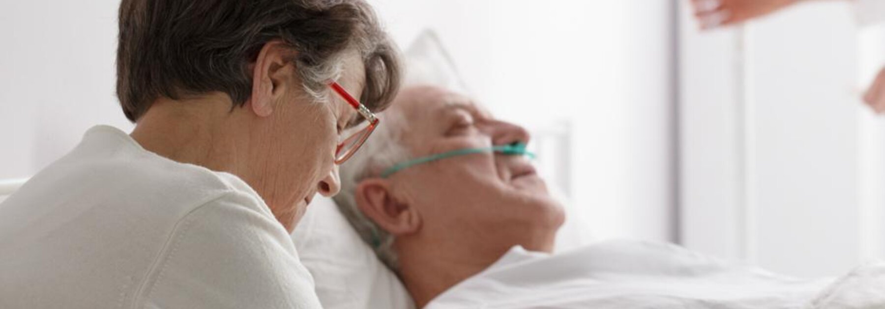 In diesem Krankenhaus sitzt eine Ehefrau bei ihrem sich erholenden Mann, während ein Arzt im Hintergrund seinen Infusionsbeutel anpasst.