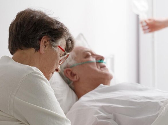 In diesem Krankenhaus sitzt eine Ehefrau bei ihrem sich erholenden Mann, während ein Arzt im Hintergrund seinen Infusionsbeutel anpasst.
