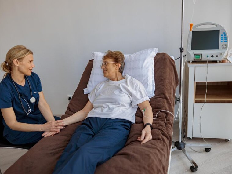 Eine Frau liegt in einem Krankenhausbett, neben ihr sitzt ein Arzt und hält ihre Hand.