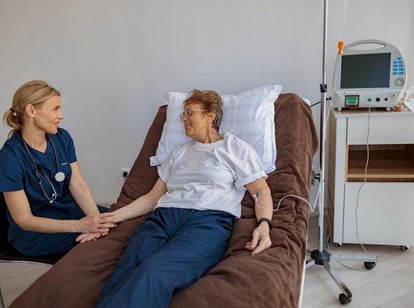 Eine Frau liegt in einem Krankenhausbett, neben ihr sitzt ein Arzt und hält ihre Hand.
