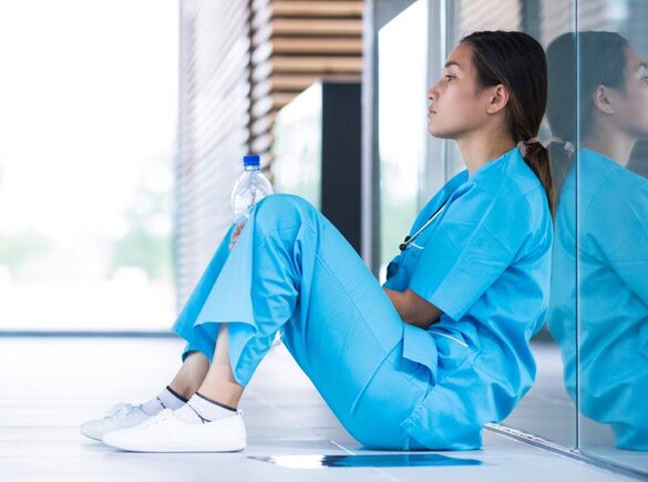 Krankenschwester sitzt auf dem Boden eines Flurs und lehnt mit dem Rücken an einer Glaswand.