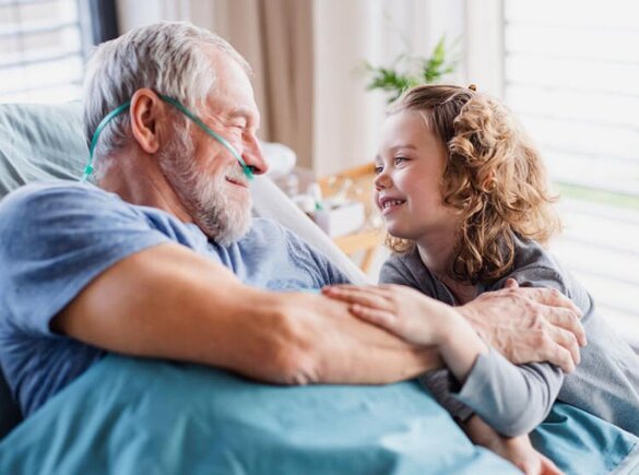 Älterer Mann, der mit einer Nasenkanüle in einem Krankenhausbett liegt und ein junges Mädchen anlächelt, das neben ihm sitzt.