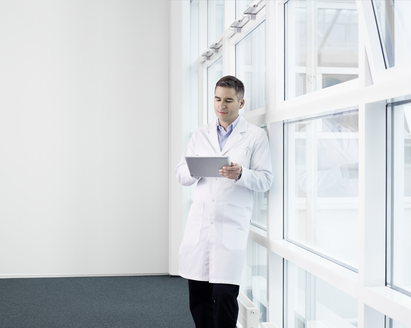 Doctor leaning against a wall of glass windows and looking at an electronic tablet