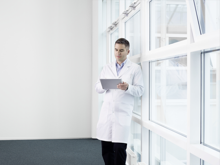 Doctor leaning against a wall of glass windows and looking at an electronic tablet