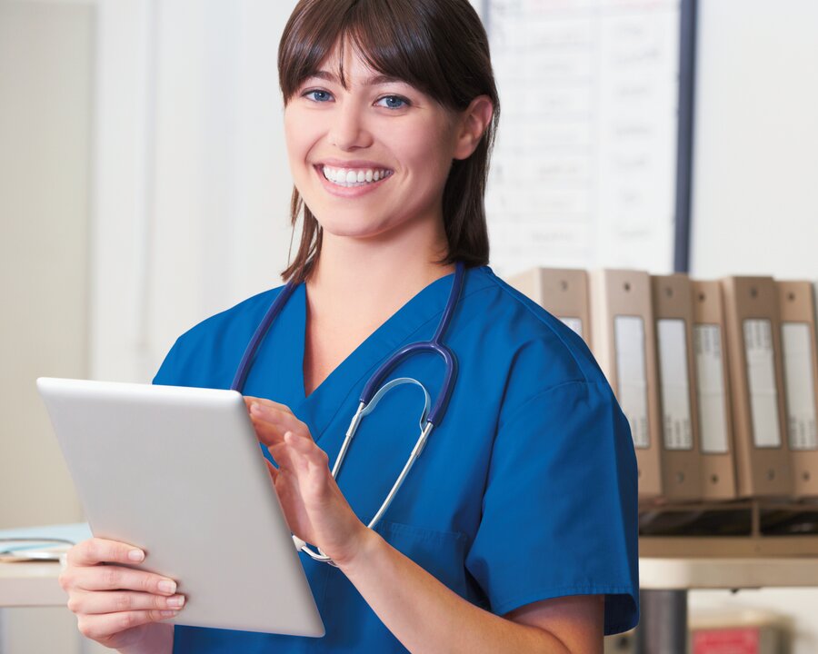 Eine medizinische Fachkraft gibt Informationen in ein Tablet ein und blickt dabei in die Kamera.