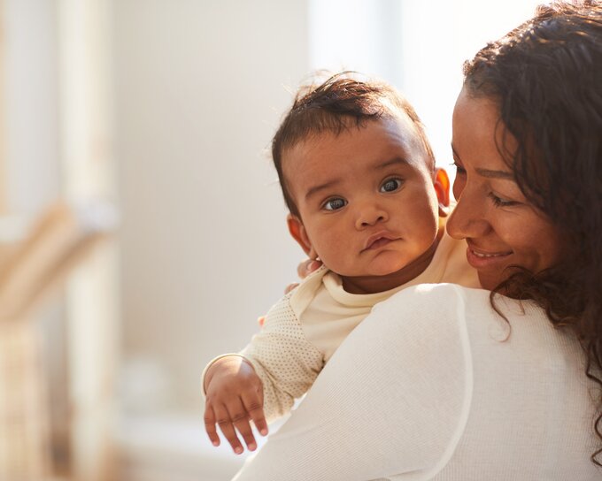 A mother holds her baby and looks at it while the baby looks at the camera, outside.