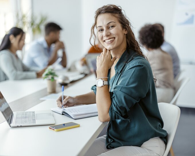 Female employee sitting at conference table writing in a notebook. In front of her is an open laptop.  Next to her is a group of other employees talking together.