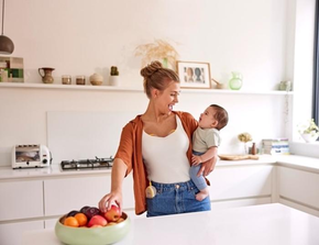 Een moeder houdt haar baby vast en eet een snack terwijl ze haar Medela Freestyle™ Hands-free dubbele elektrische borstkolf draagt.
