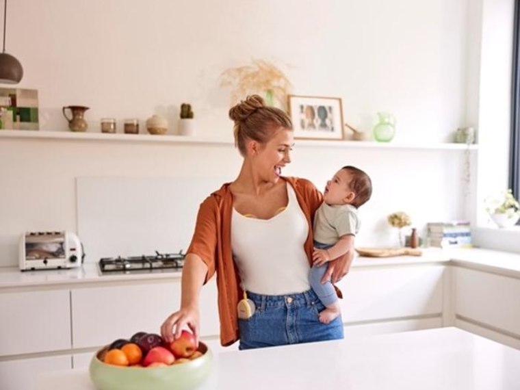 Eine Mutter hält ihr Baby und isst einen Snack, während sie ihre Medela Freestyle™ Hands-free elektrische Doppelmilchpumpe trägt.