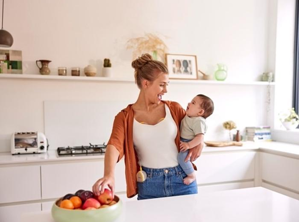 A mom holds her baby and has a snack while wearing her Medela Freestyle™ Hands-free double electric breast pump.