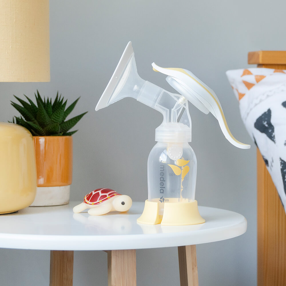 A Medela Harmony™ manual breast pump on a bedside table with a lamp, a plant and a toy.