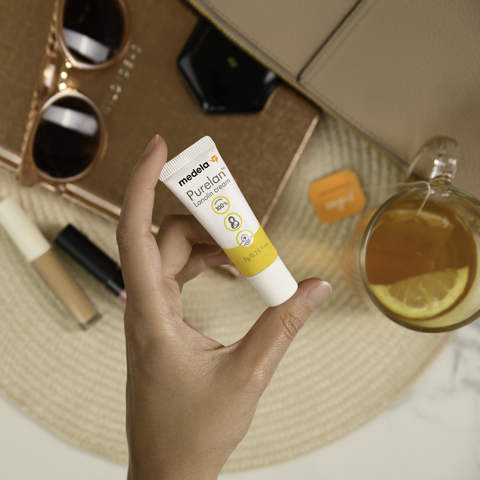 Eine Person hält eine 7-g-Tube Medela Purelan™ Lanolincreme in der Hand über Sonnenbrille, Make-up und Tee für unterwegs.