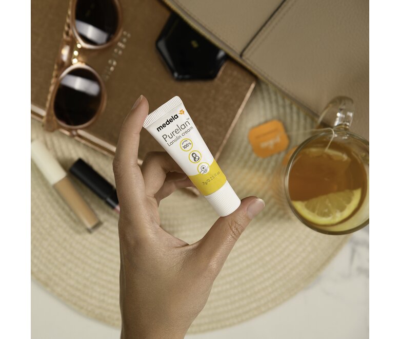 Eine Person hält eine 7-g-Tube Medela Purelan™ Lanolincreme in der Hand über Sonnenbrille, Make-up und Tee für unterwegs.