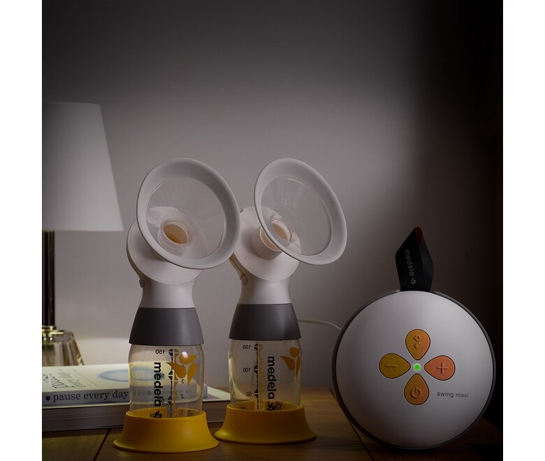 Die elektrische Doppelmilchpumpe Medela Swing Maxi™ mit zwei Classic-Auffangbehältern auf einem Nachttisch mit gedämpftem Licht.