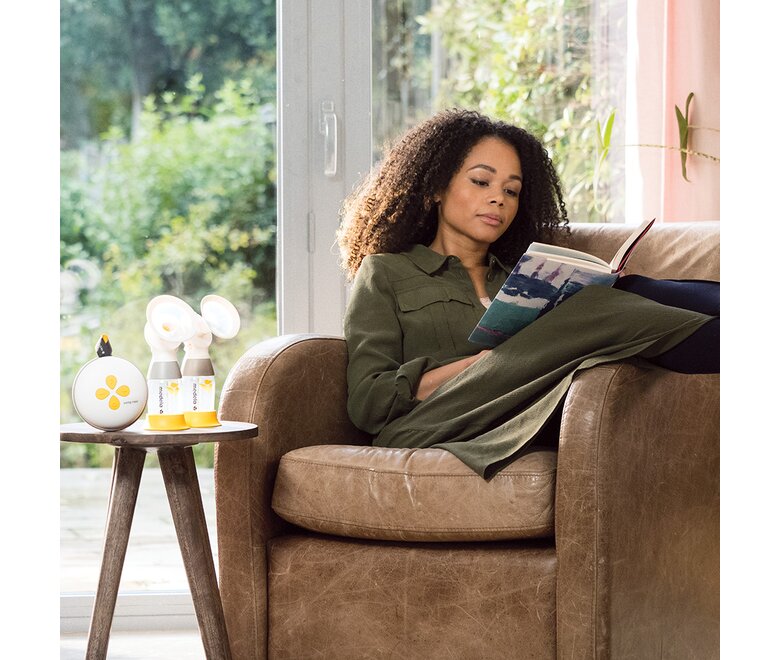 Eine Frau, die sich zu Hause entspannt und liest, mit ihrer Medela Swing Maxi™ Hands-free Electric Breast Pump und klassischen Sammelbehältern neben sich.