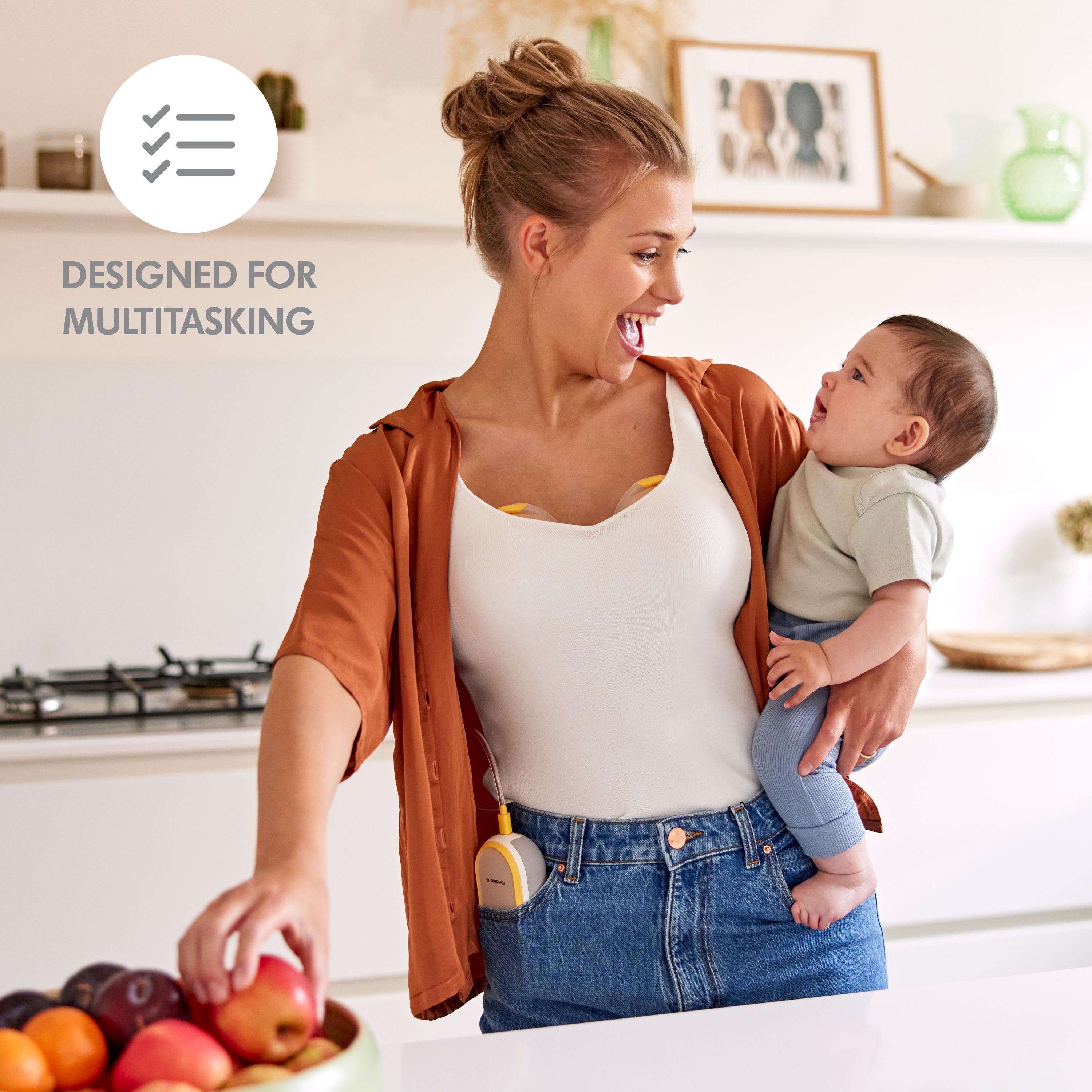 A mother holds her baby and has a snack while wearing her Medela Freestyle™ Hands-free double electric breast pump. Text: “Designed for multitasking”