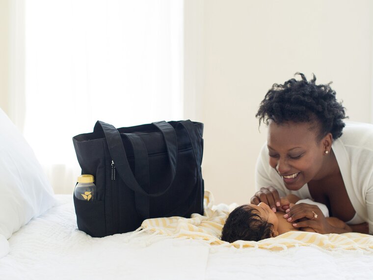 Baby laying on bed next to a Medela pumping bag.  Woman is smiling down on baby.