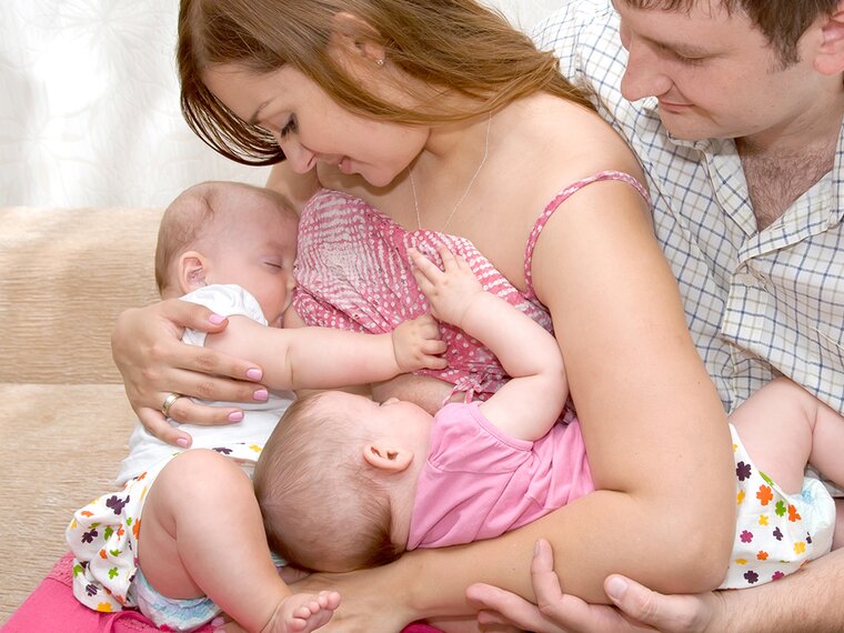 Een moeder voedt haar tweeling in tandem terwijl de vader naast hen zit en een van de baby's ondersteunt.