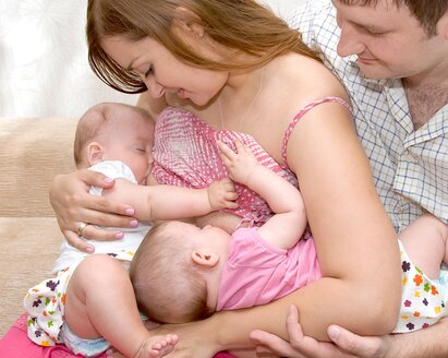 A mom feeds her twins in tandem while the dad sits next to them and supports one of the babies.