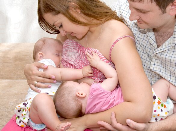 A mom feeds her twins in tandem while the dad sits next to them and supports one of the babies.