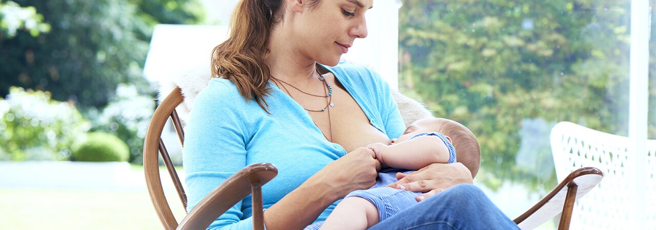 Une mère allaite son bébé, assise dans un fauteuil à bascule près de la fenêtre.