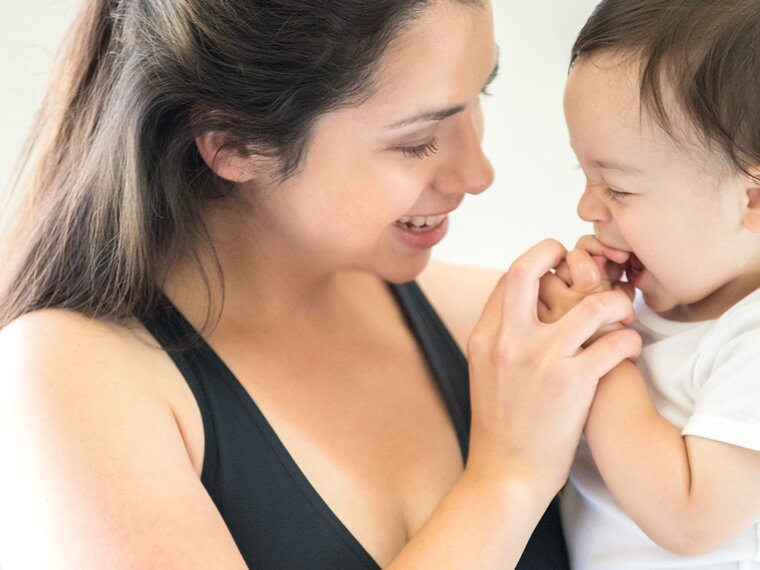 Een baby probeert zijn handjes in zijn mond te stoppen en zijn moeder houdt speels zijn handjes vast, waardoor ze glimlachen.
