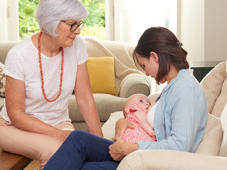 A mother breastfeeds her baby at home in the living room while the baby's grandmother supports her.