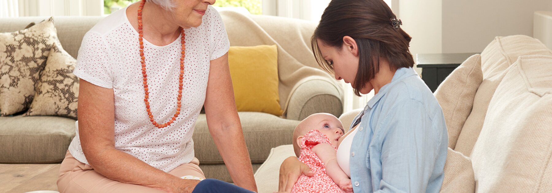 A mother breastfeeds her baby at home in the living room while the baby's grandmother supports her.