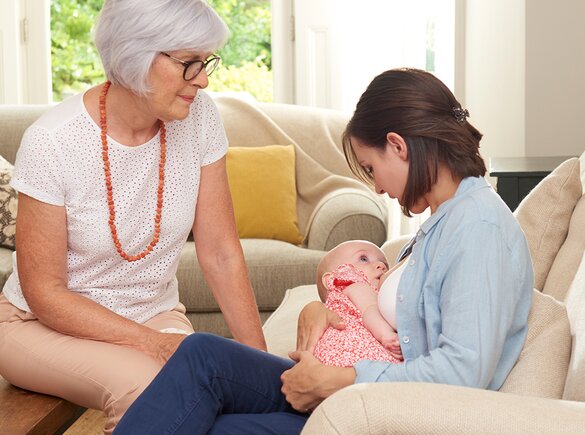 Eine Mutter stillt ihr Kind zu Hause im Wohnzimmer, während die Großmutter des Kindes sie unterstützt.