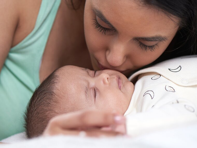 Baby lying on a bed and mother leaning over baby and kissing baby's cheek