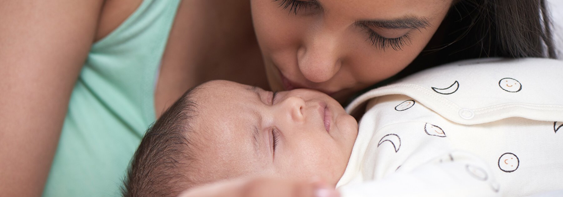 Baby lying on a bed and mother leaning over baby and kissing baby's cheek