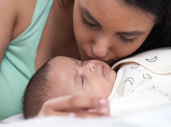 Baby lying on a bed and mother leaning over baby and kissing baby's cheek