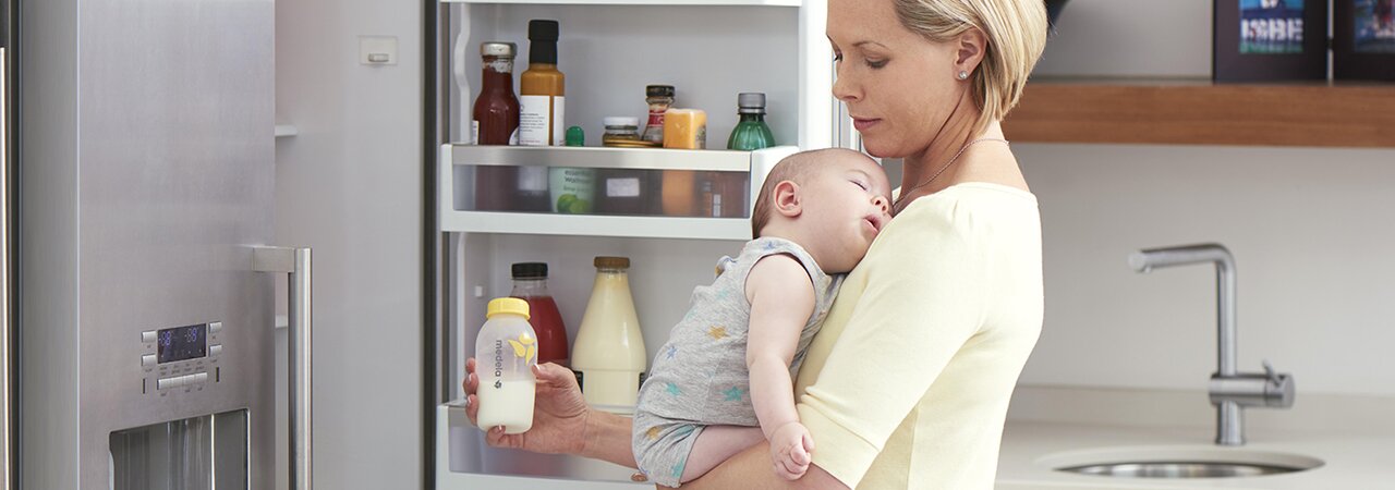 Una mamma tiene in braccio il suo bambino in cucina mentre prepara il latte espresso refrigerato per la poppata.