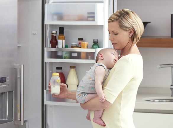 Eine Mutter hält ihr Baby in der Küche, während sie gekühlte Muttermilch für eine Fütterung vorbereitet.