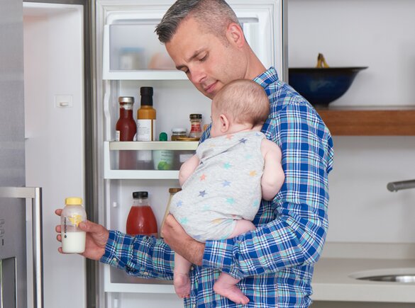 Um pai segura o seu bebé na cozinha enquanto tira um biberão do frigorífico para o aquecer para a alimentação.