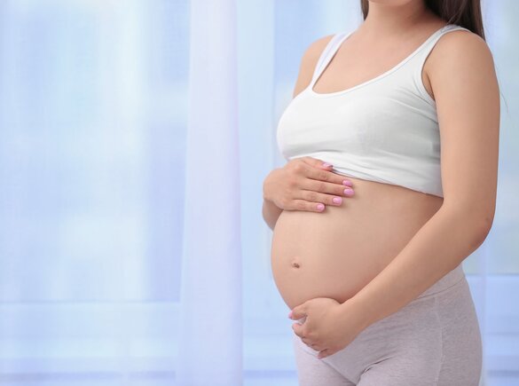 Eine schwangere Frau hält im Stehen ihren unbedeckten schwangeren Bauch.