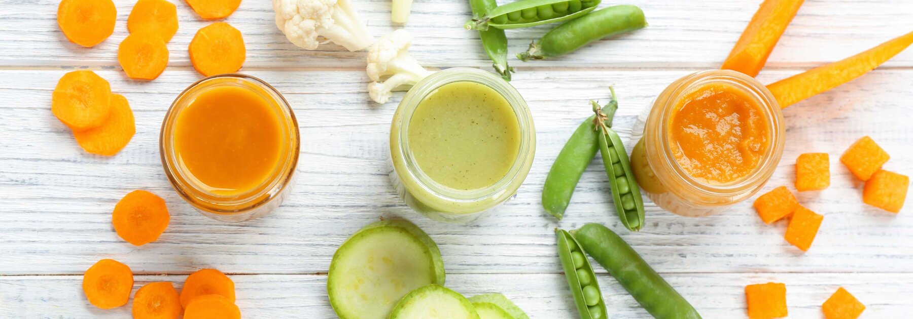 Ein Tisch mit drei gefüllten Gläsern mit Babynahrung, umgeben von geschnittenem Gemüse: Karotten, Erbsen, Blumenkohl, Gurken und Kürbis.