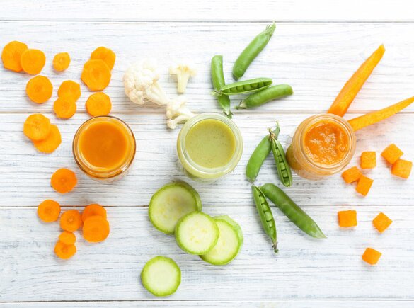 Ein Tisch mit drei gefüllten Gläsern mit Babynahrung, umgeben von geschnittenem Gemüse: Karotten, Erbsen, Blumenkohl, Gurken und Kürbis.
